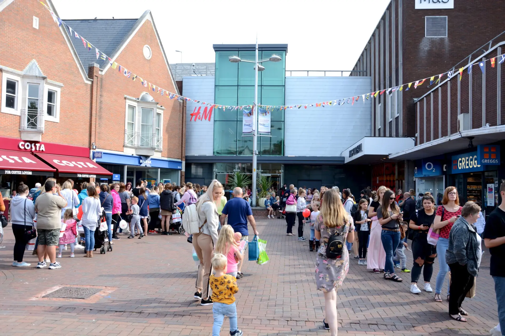 Image of Falkland Square, Poole: A bustling scene with numerous people gathered in the square. On the left, there is a Costa Coffee shop with a crowd outside. Nearby, a Halifax bank is visible. In the center, there's a prominent H&M store with people milling about. On the right side, there are shops including Greggs and O2. Colorful bunting stretches across the square above the crowd, adding a festive atmosphere. People of all ages, including families with children, are seen enjoying the day, some carrying shopping bags or pushing strollers.