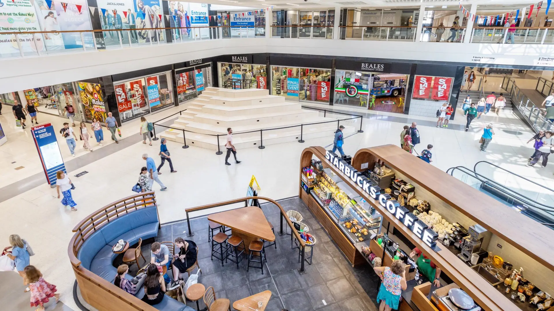 Image of The Dolphin Poole interior: A bright and spacious indoor shopping center with multiple levels. In the foreground, a Starbucks Coffee kiosk with patrons seated around it is visible. Shoppers are walking along the wide walkways on both levels, with various stores including Beales displaying large sale signs. An open stairway and escalator are seen on the right, facilitating movement between floors. Colorful banners and advertisements add to the lively atmosphere. The scene captures a mix of people shopping, socializing, and enjoying their time in the mall.