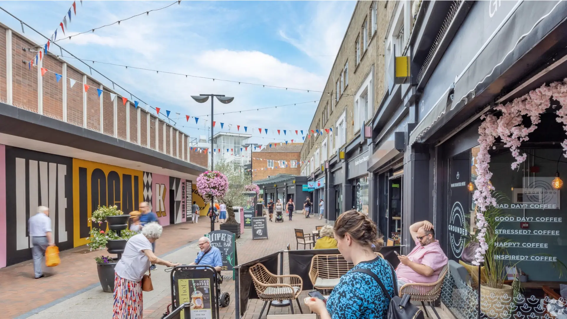 A lively street scene at Kingland Crescent, Poole, featuring a pedestrian area lined with shops and outdoor seating. People are enjoying the pleasant weather, with some sitting at tables outside a café named "Grounded Coffee" adorned with pink floral decorations. The café's window displays a humorous sign offering coffee for various moods: "Bad day? Coffee," "Good day? Coffee," "Stressed? Coffee," "Happy? Coffee," and "Inspired? Coffee." Colorful bunting is strung across the street, adding to the festive atmosphere. In the background, a large mural with the words "LIVE LOCAL" enhances the vibrant and community-focused ambiance. Potted flowers and greenery contribute to the inviting and cheerful environment.