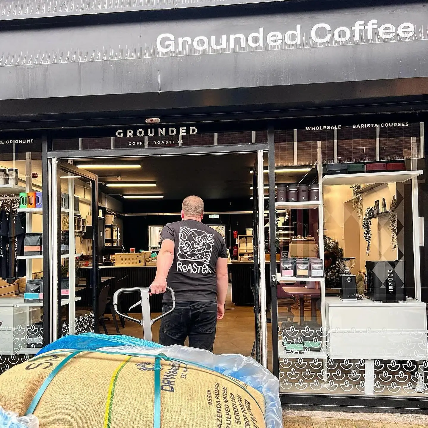Exterior of Grounded Coffee cafe shopfront, a staff member is pulling a trolley with a large burlap sack full of coffee beans into the front door