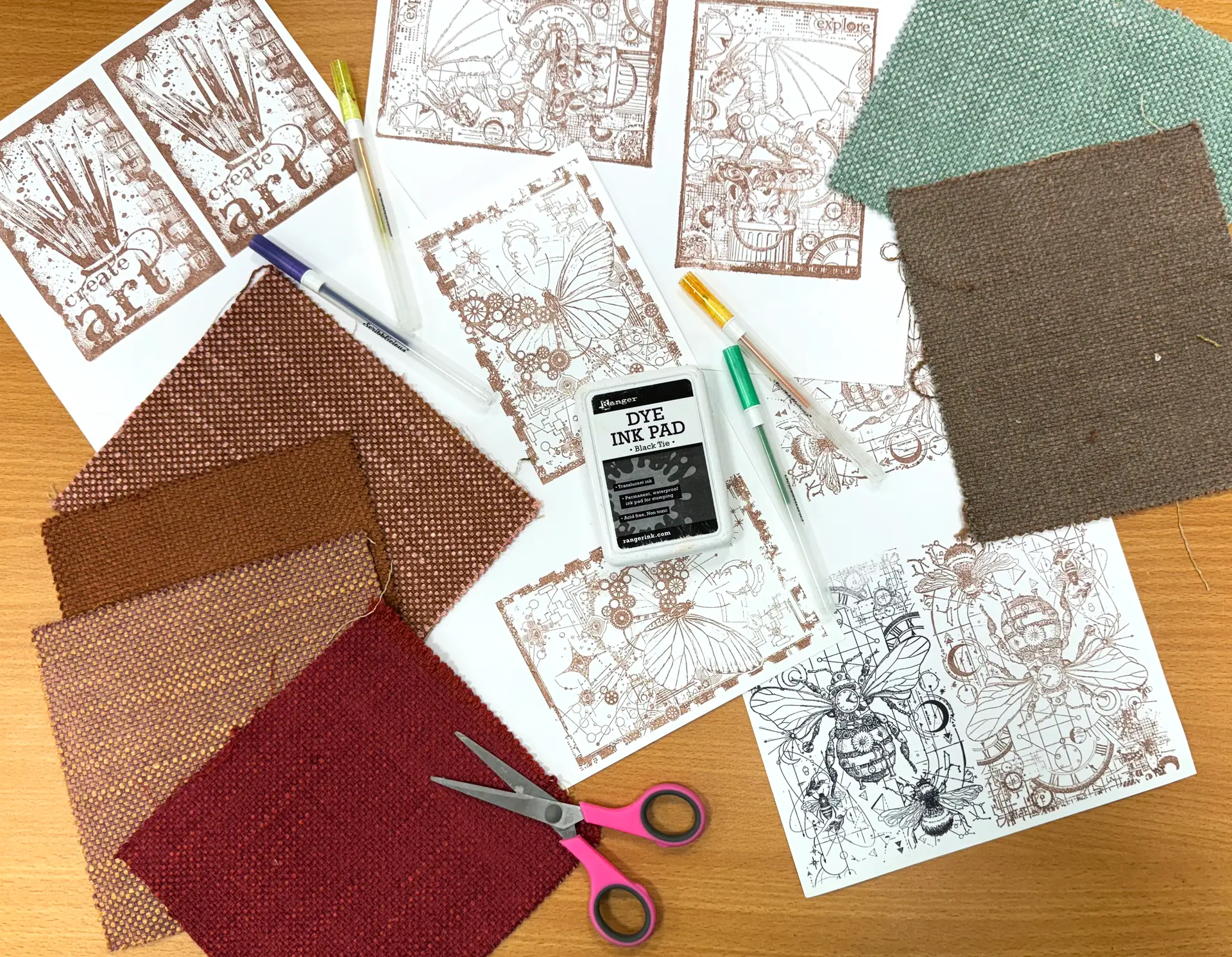 A selection of art and craft materials on a wooden tabletop. Items include squares of fabric, gel pens, craft scissors and intricate ink print designs.