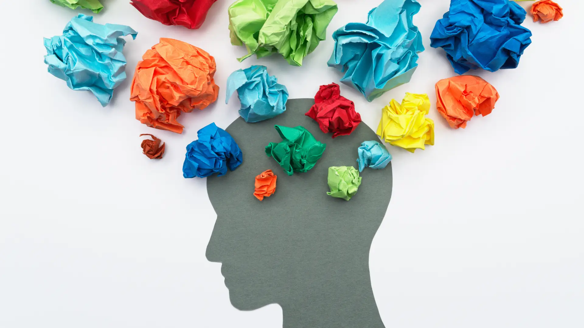 A grey silhouette of a head in side profile against a white background. Brightly coloured pieces of paper of various sizes are scrunched up and arranged around the top of the head to represent thoughts or worries.