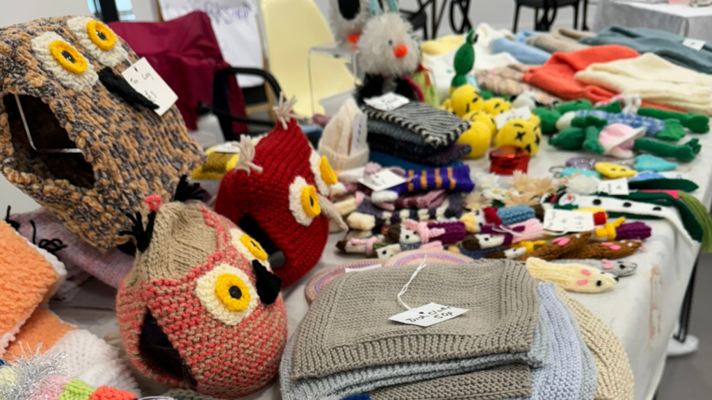 A selection of handmade, crocheted and knitted items arranged for sale at an indoor market. Including crocheted owls, finger puppets and children's clothing.