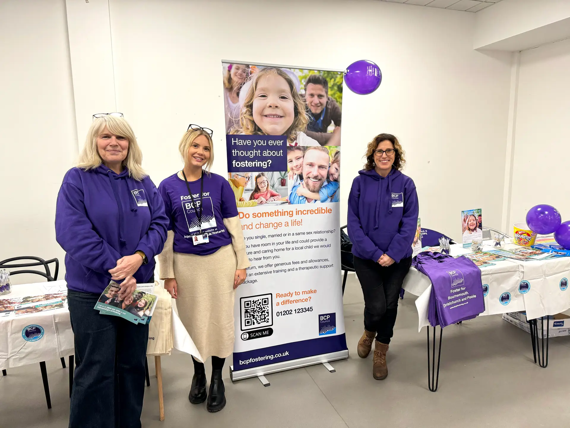 Three members of staff from BCP Council's Fostering and Supported Lodgings team at a previous information event. They stand beside tables with promotional leaflets and merchandise, as well as a roller banner which says 'Have you ever thought about fostering? Do something incredible and change a life!'