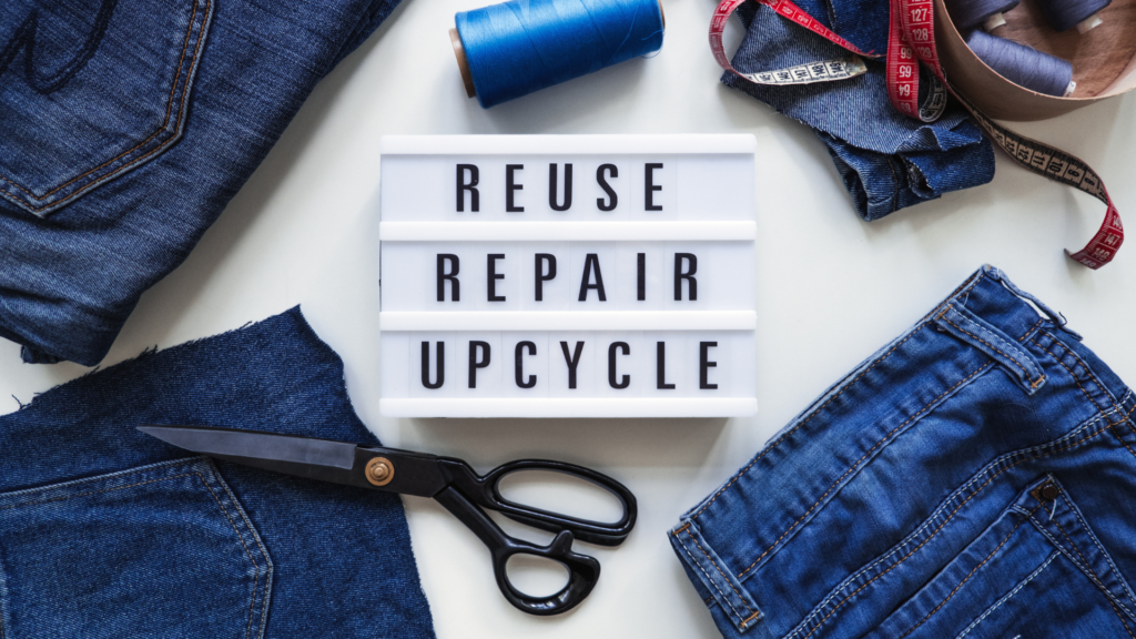 A white box sign reading, 'Reuse, Repair, Recycle,' surrounded by denim fabric, dressmaking scissors and spools of thread.