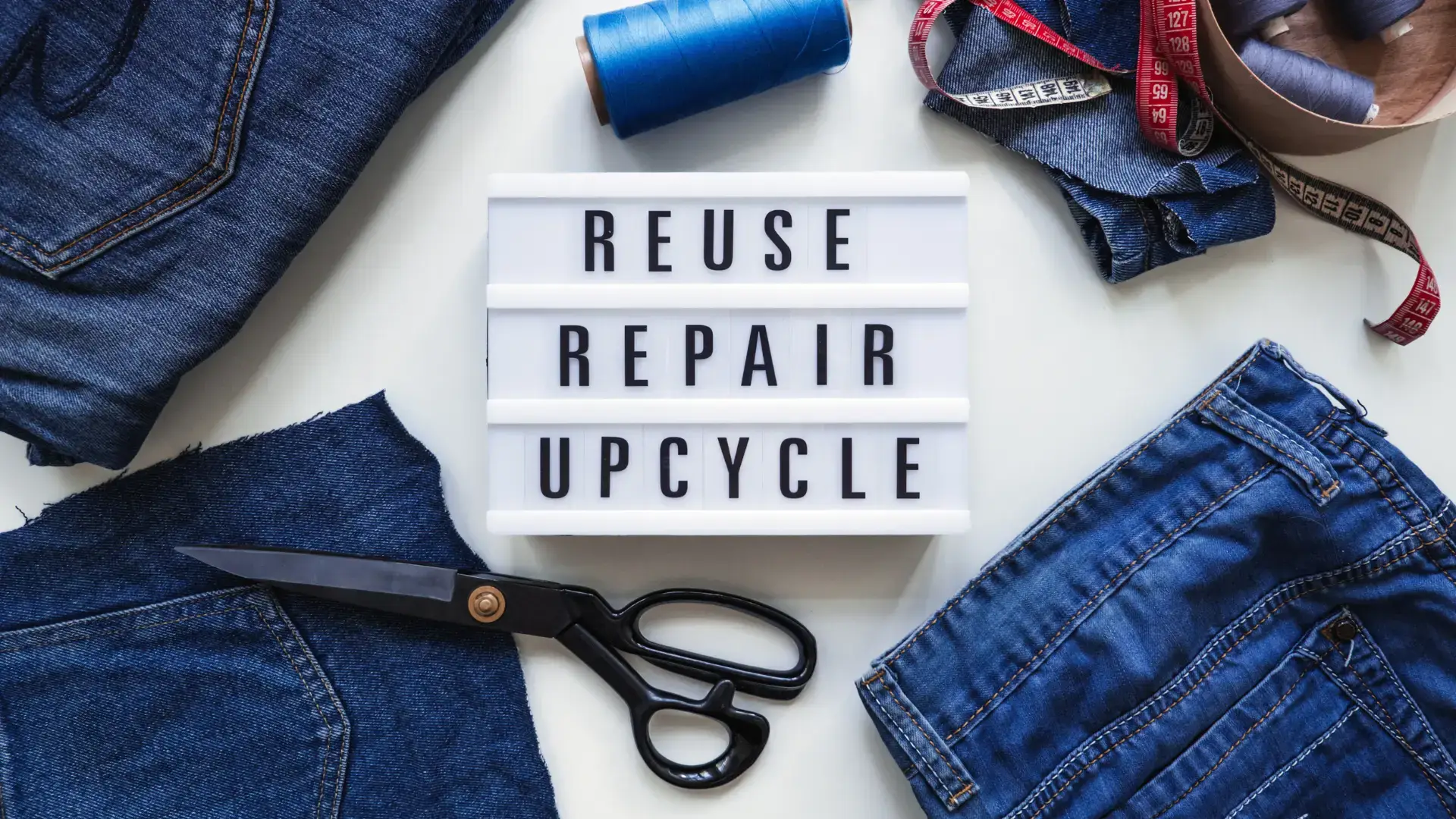 A white box sign reading, 'Reuse, Repair, Recycle,' surrounded by denim fabric, dressmaking scissors and spools of thread.