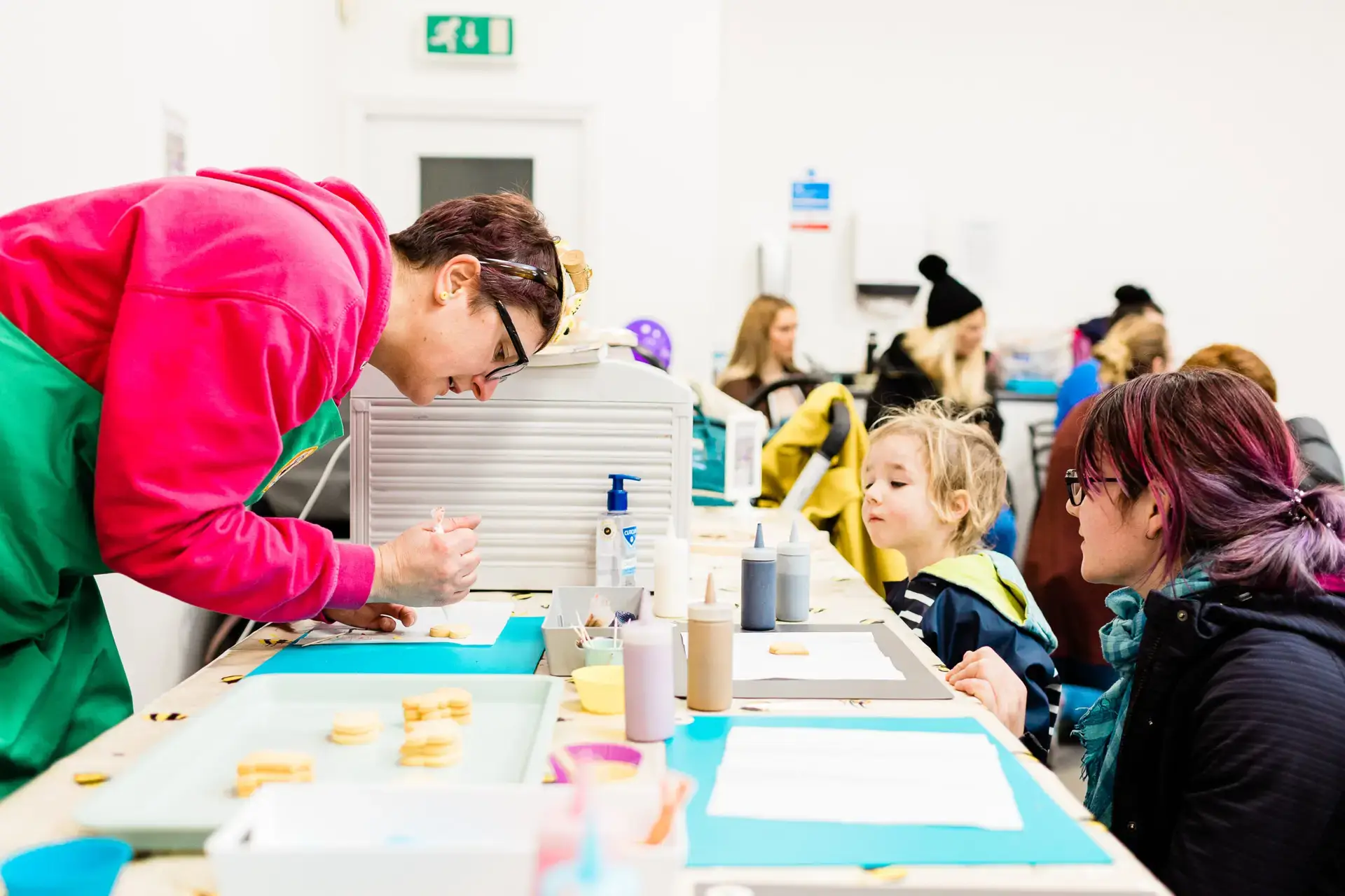 A woman decorating a sugar biscuit with brightly coloured icing. A small boy and his mother watch with interest.
