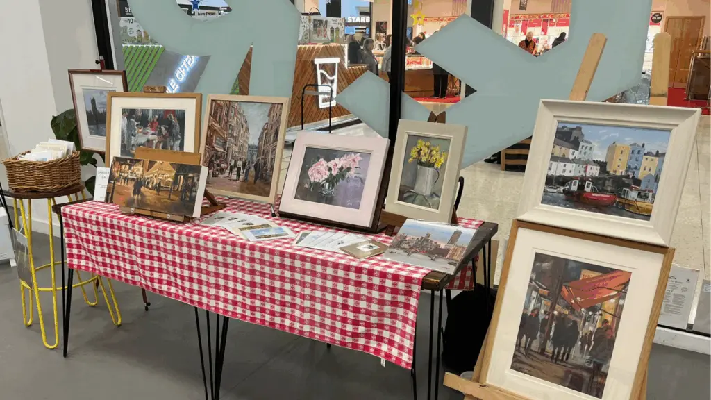 A display table covered with a red and white checkered tablecloth showcases various framed paintings. The table is set up inside The Dolphin's GATHER event space. There are eight framed artworks on the table and an easel next to it holding two more paintings. The paintings depict various scenes including flowers, cityscapes, and market scenes. A wicker basket with napkins or papers is placed on a small yellow stool to the left of the table. In the background, there are large glass windows showing a busy indoor area with shops and people.