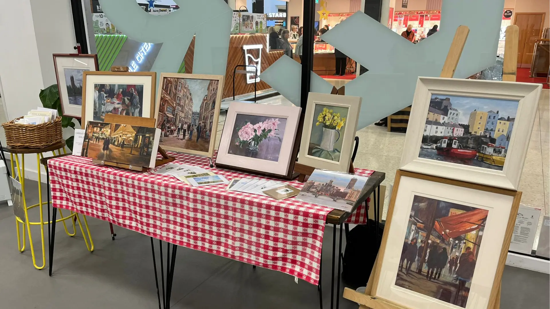 A display table covered with a red and white checkered tablecloth showcases various framed paintings. The table is set up inside The Dolphin's GATHER event space. There are eight framed artworks on the table and an easel next to it holding two more paintings. The paintings depict various scenes including flowers, cityscapes, and market scenes. A wicker basket with napkins or papers is placed on a small yellow stool to the left of the table. In the background, there are large glass windows showing a busy indoor area with shops and people.